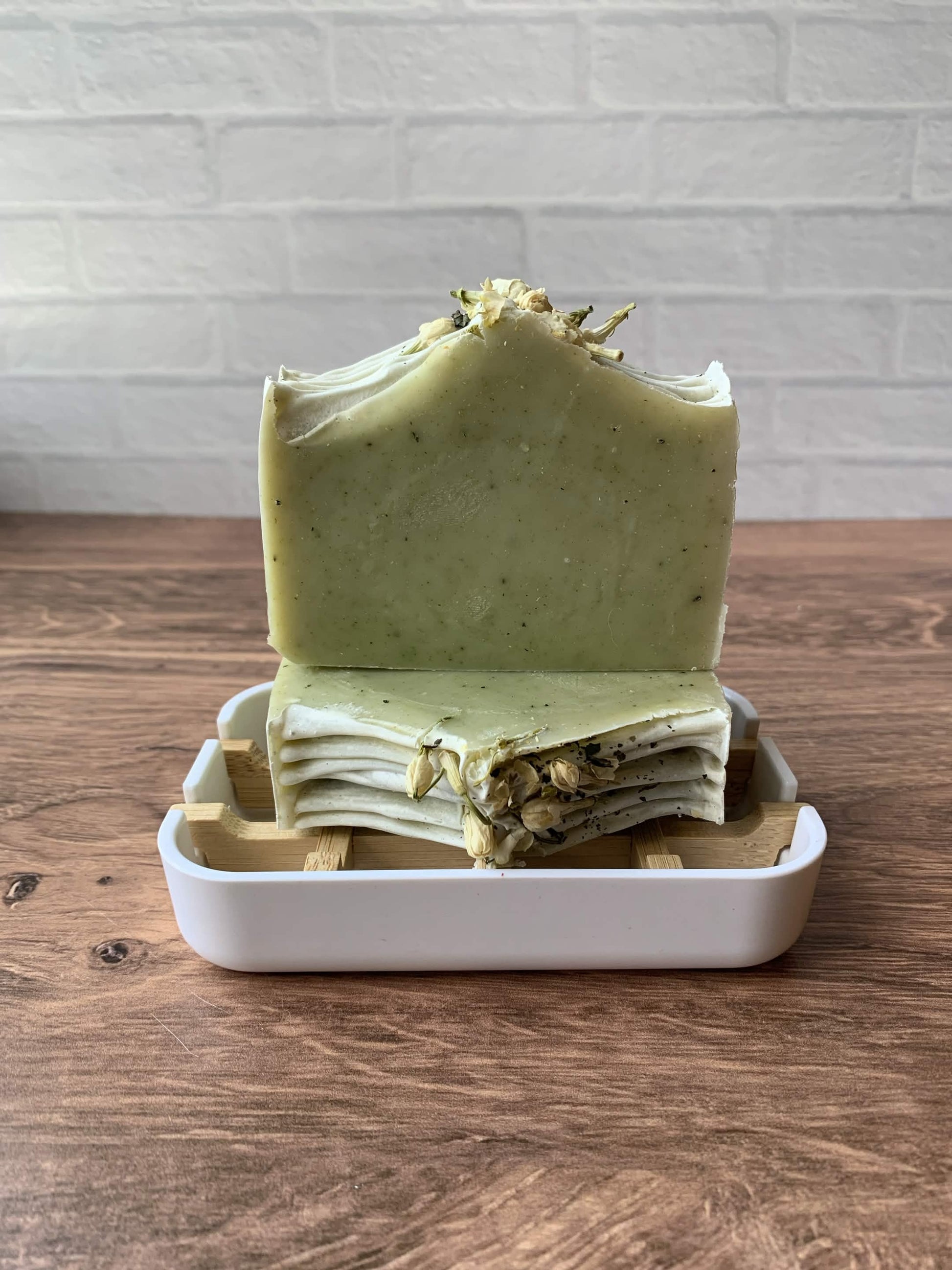 A handmade bar of pale green speckled soap stands upright in a white rectangular soap dish with a wooden slotted tray behind it, set on a wooden tabletop against a white brick wall.